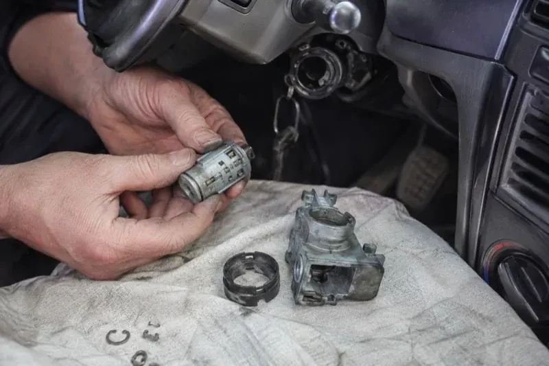 Hands holding a removed ignition cylinder beside a car steering column, with the housing and small parts on a cloth. — Ignition Lock Repair, Mississauga